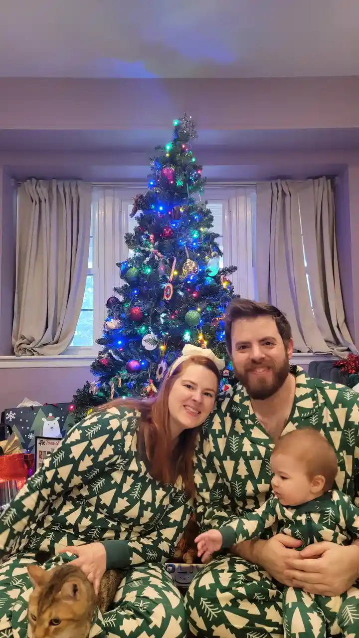A family of three — a man, a woman, and a baby — are sitting together in matching green pajamas with white tree patterns…