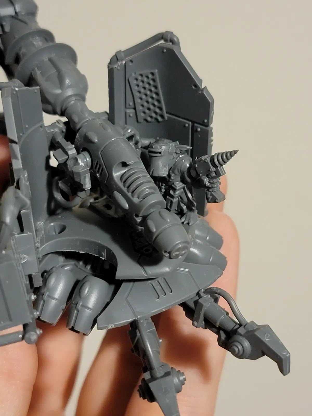 A close-up photo shows a person's hand holding an unpainted, grey plastic Warhammer 40k miniature—a heavily armed, armored…
