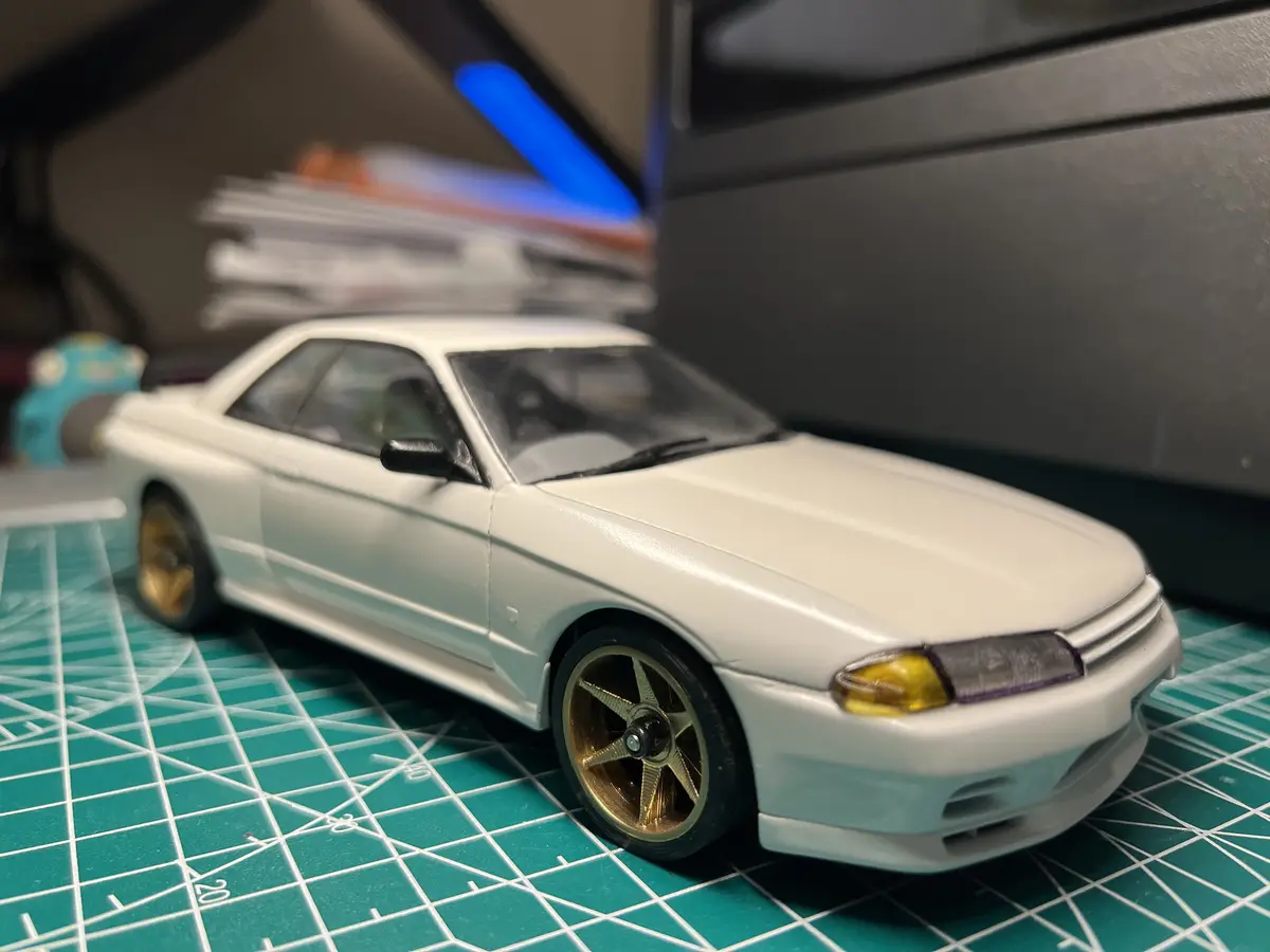 A white Nissan Skyline R32 model car with gold wheels sits on a green cutting mat with grid lines. The background is…