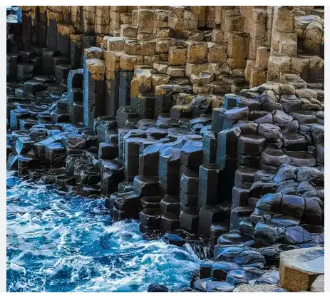 The image shows a natural rock formation with hexagonal basalt columns rising from the ocean, with waves crashing around…