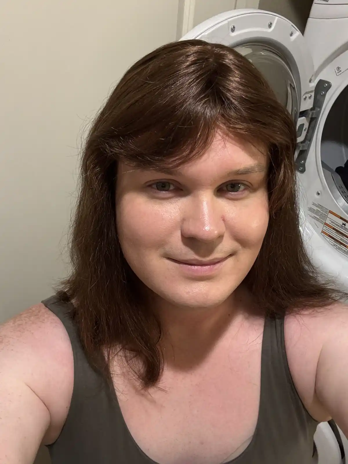 A person with shoulder-length brown hair and a subtle smile takes a selfie in front of an open white front-loading washing…