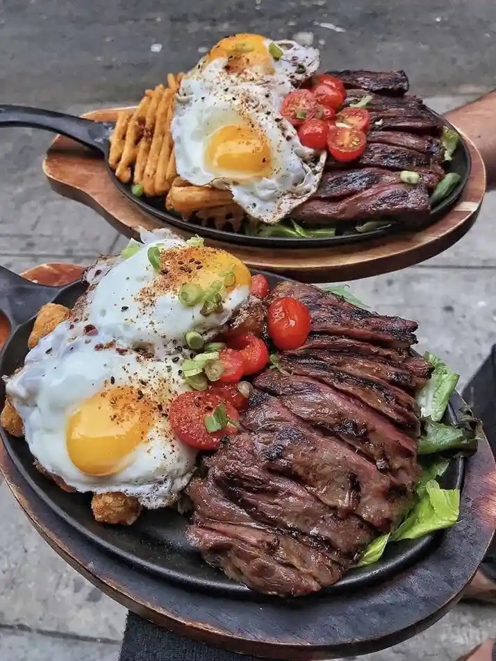 Two identical black cast-iron skillet meals are stacked, each featuring sliced grilled steak, two sunny-side-up eggs with…