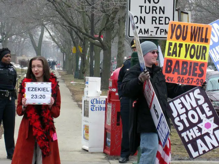 A man holds multiple antisemitic signs including 'YOU WILL EAT YOUR BABIES' and 'JEWS STOLE THE LAND', while a woman holds a…