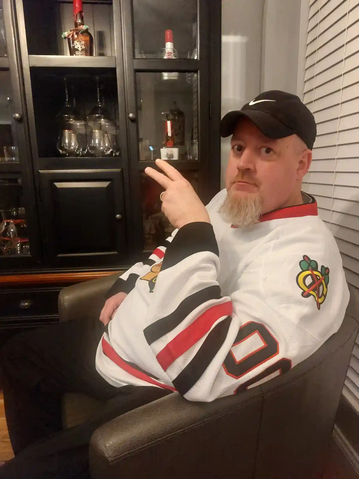 a man with a goatee and black cap sits in a leather chair, wearing a white Chicago Blackhawks hockey jersey with the number…