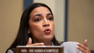 A woman with dark hair and a surprised expression is speaking at a hearing, her hands gesturing as she talks. A nameplate in…