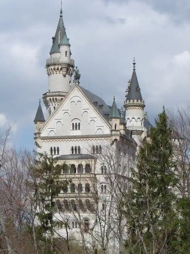 A photograph of the Neuschwanstein Castle in Germany, featuring its iconic white stone towers and green roofs, partially…