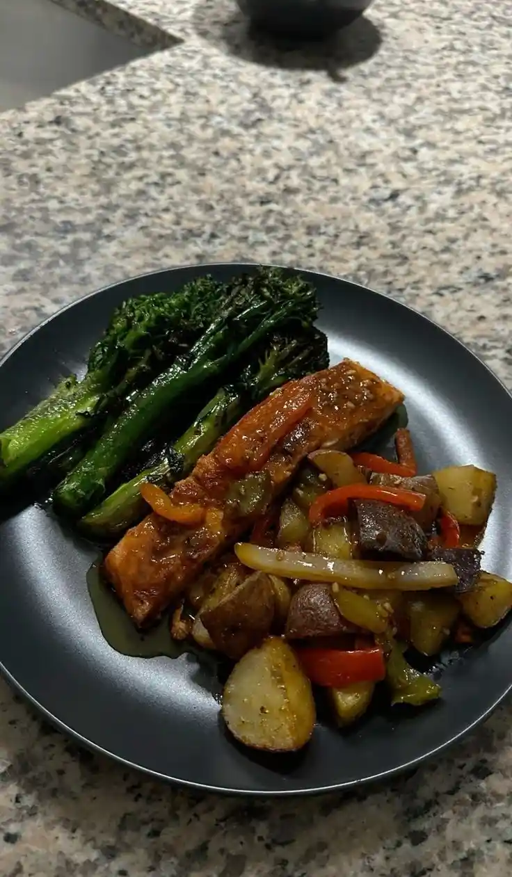 A close-up photo of a plated meal featuring grilled salmon, roasted broccolini, and a colorful medley of roasted potatoes…