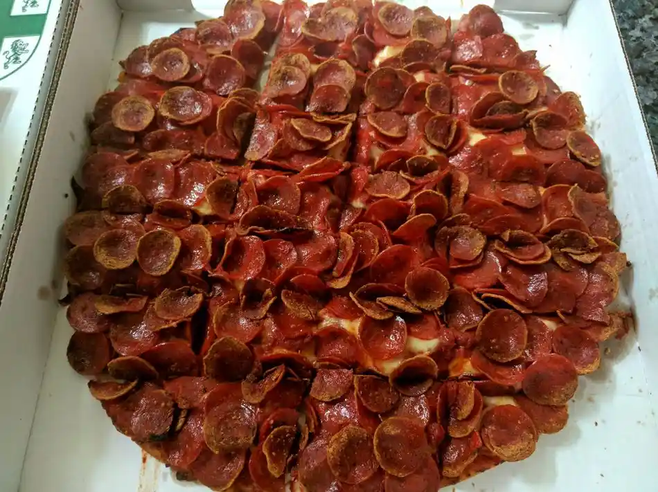 A close-up photo of a pepperoni pizza in a white cardboard box, where the pepperoni slices are so densely packed they…