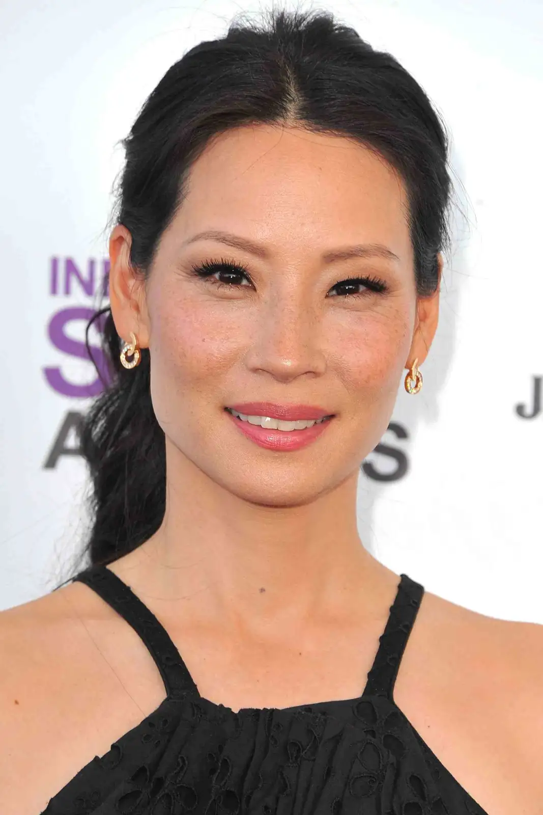 A close-up photo of a woman with dark hair pulled back, wearing a black top and gold hoop earrings, smiling slightly at the…
