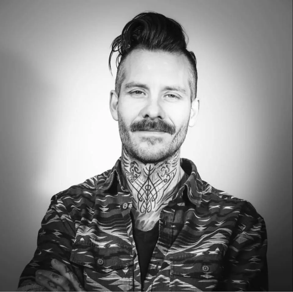 A black-and-white portrait of a man with a mustache, goatee, and intricate tattoos covering his neck and visible parts of…
