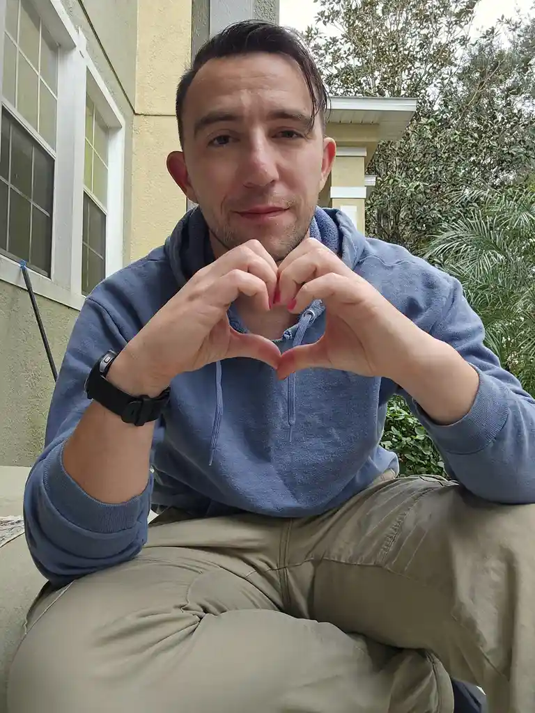A man in a blue hoodie sits outside on a porch, forming a heart shape with his hands in front of his chest. He has pink nail…