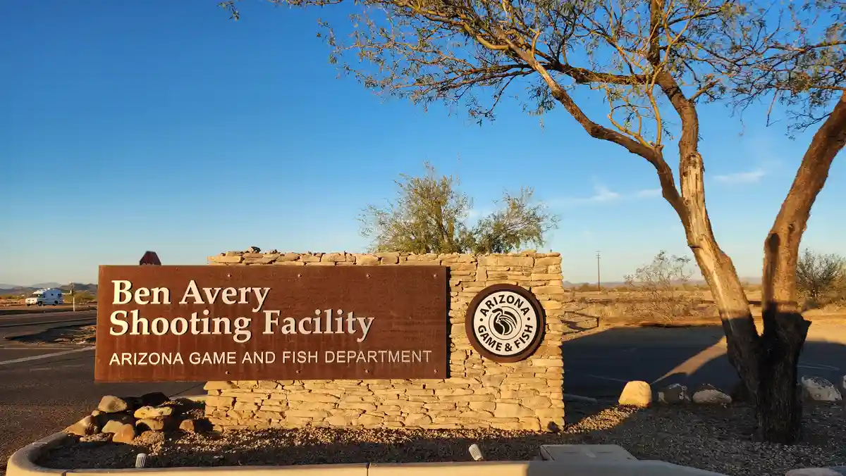 A brown sign mounted on a stone wall reads 'Ben Avery Shooting Facility' and 'ARIZONA GAME AND FISH DEPARTMENT', with a…