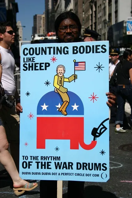 A person holds a protest sign with a cartoon of a man in a yellow suit riding a red elephant, holding an American flag. The…