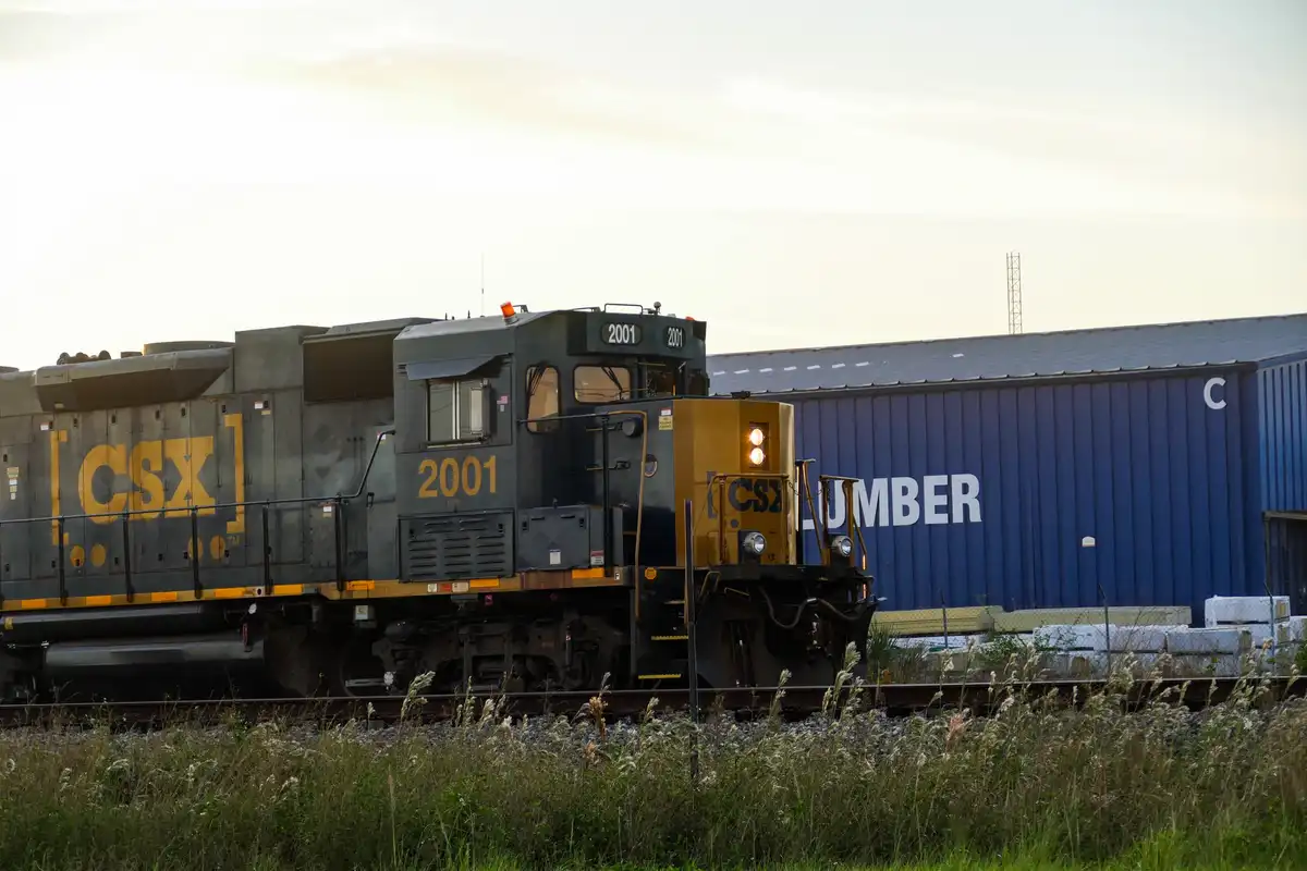 A CSX freight train locomotive numbered 2001 is shown on tracks near a blue building with 'LUMBER' written on it. The…