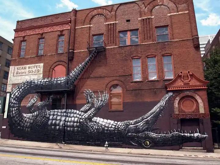 A large black-and-white mural of a giant alligator painted on the side of a red brick building, with its body stretching…