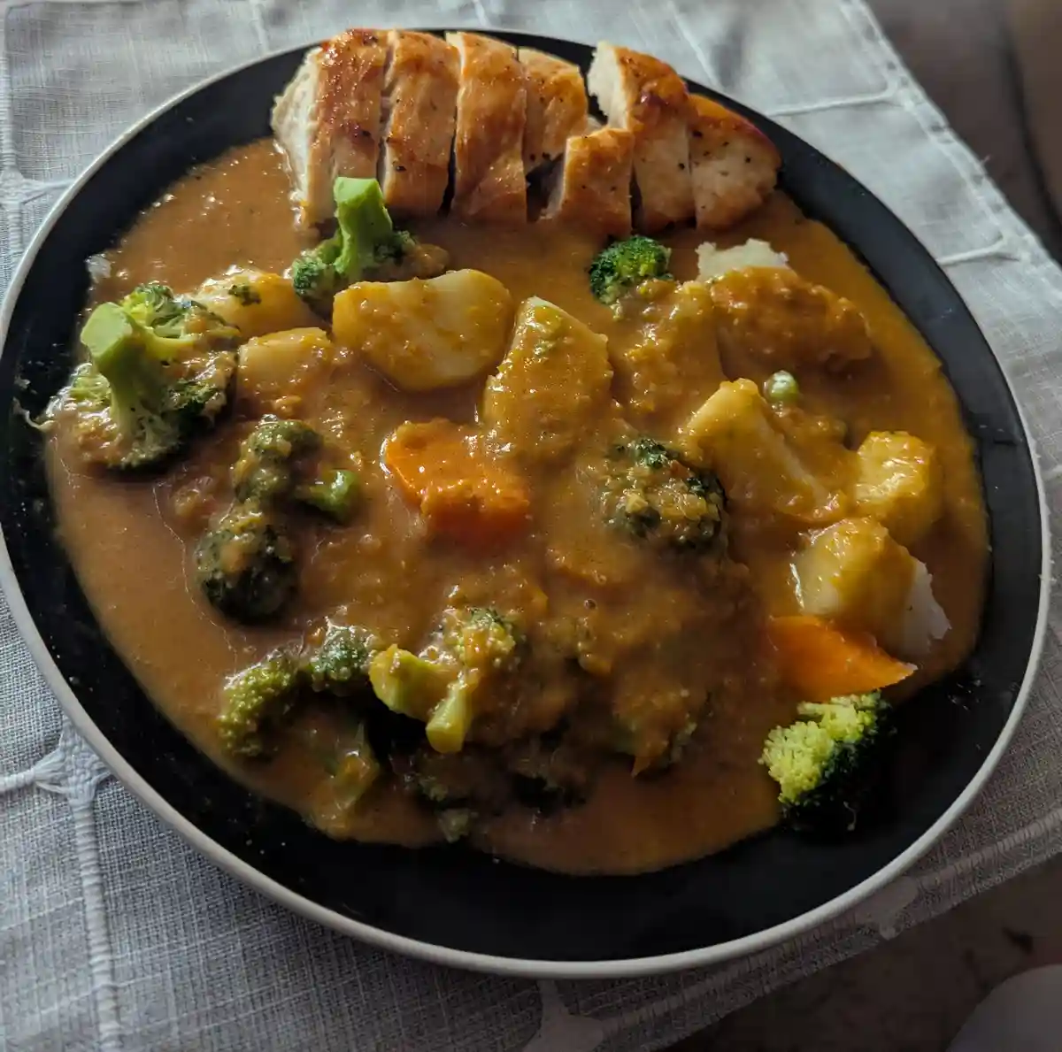 A black bowl filled with sliced chicken breast, broccoli florets, and chunks of potato or cauliflower submerged in a thick…