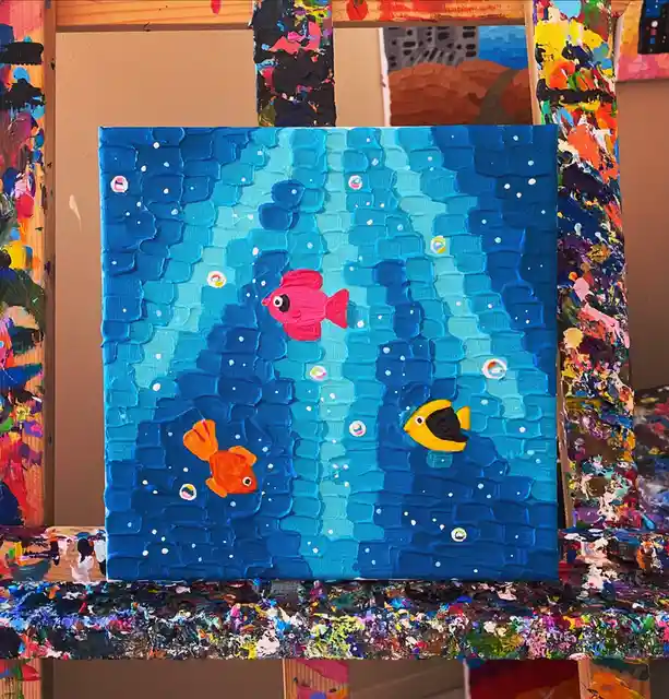 A thickly textured, colorful painting of three cartoon-style fish swimming in blue water with bubbles, displayed on an easel…
