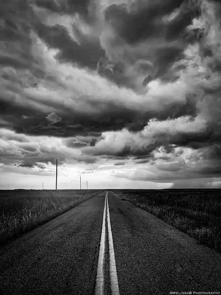 A black-and-white photograph shows a long, straight road stretching into the horizon under a dramatic, cloudy sky. The road…