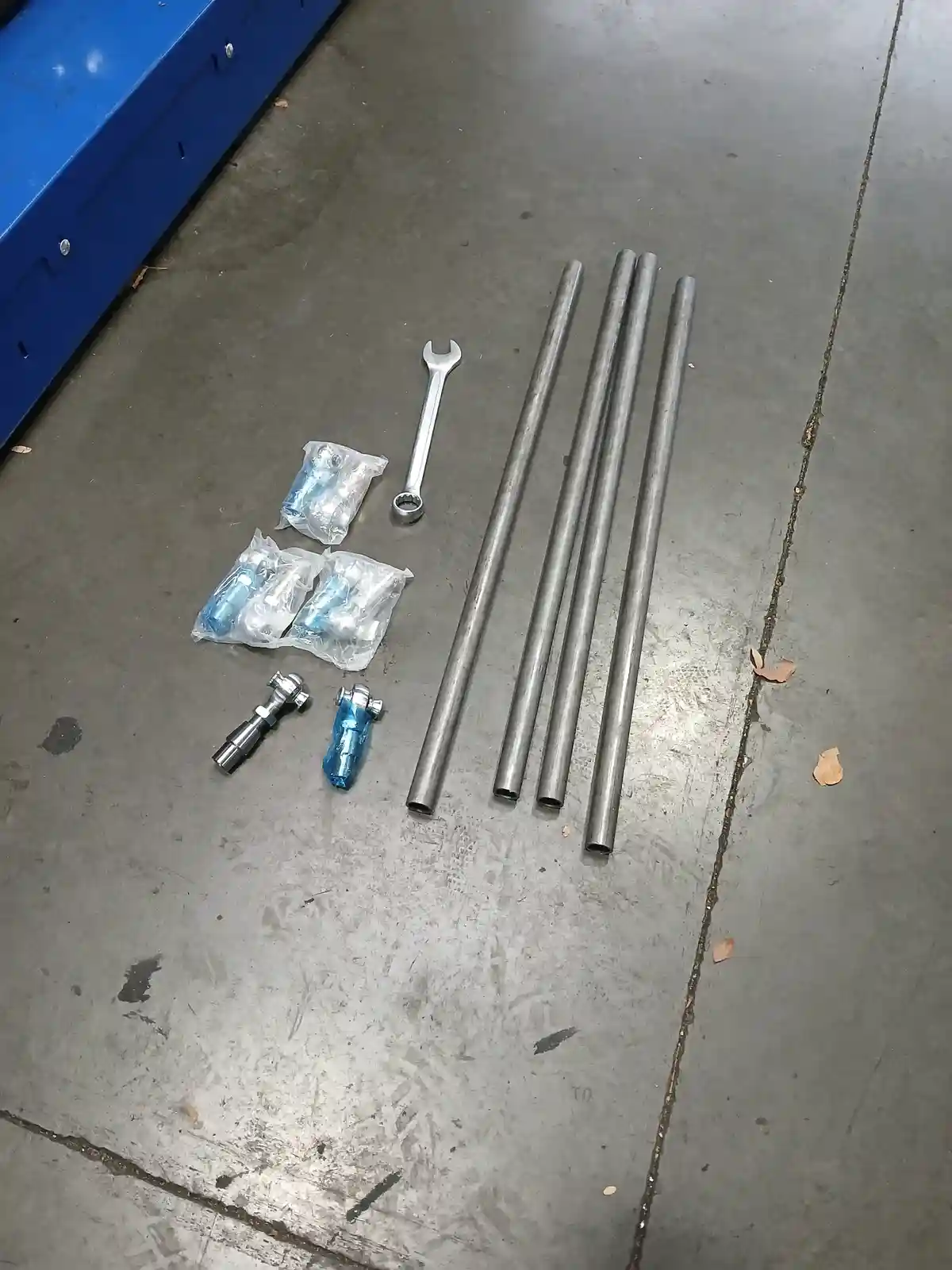 A top-down photo shows metal pipes, a wrench, and small mechanical parts on a concrete floor, with overlaid text reading 'i…