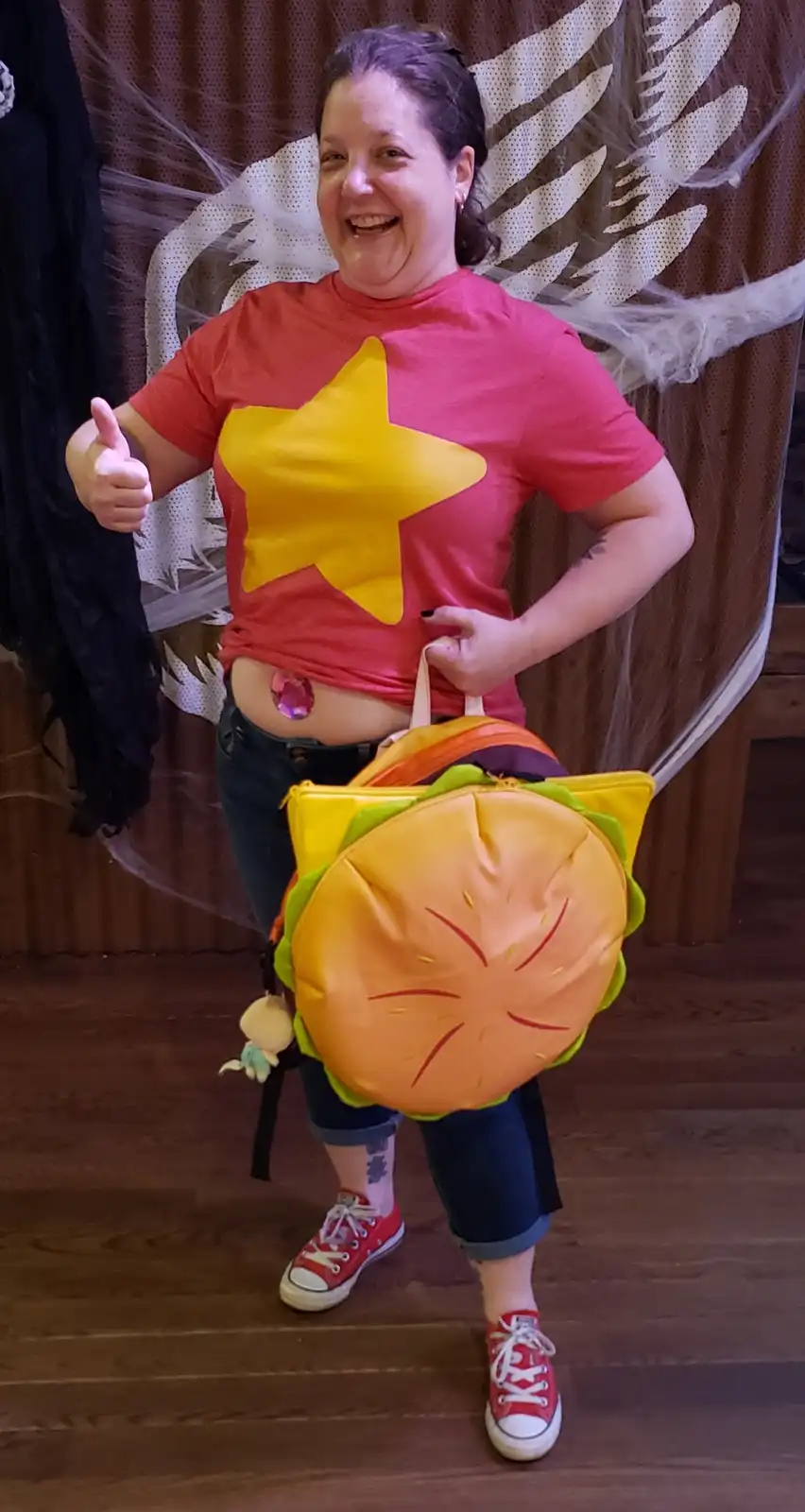 A smiling woman in a red t-shirt with a large yellow star on it, wearing angel wings and a burger-shaped backpack, gives a…