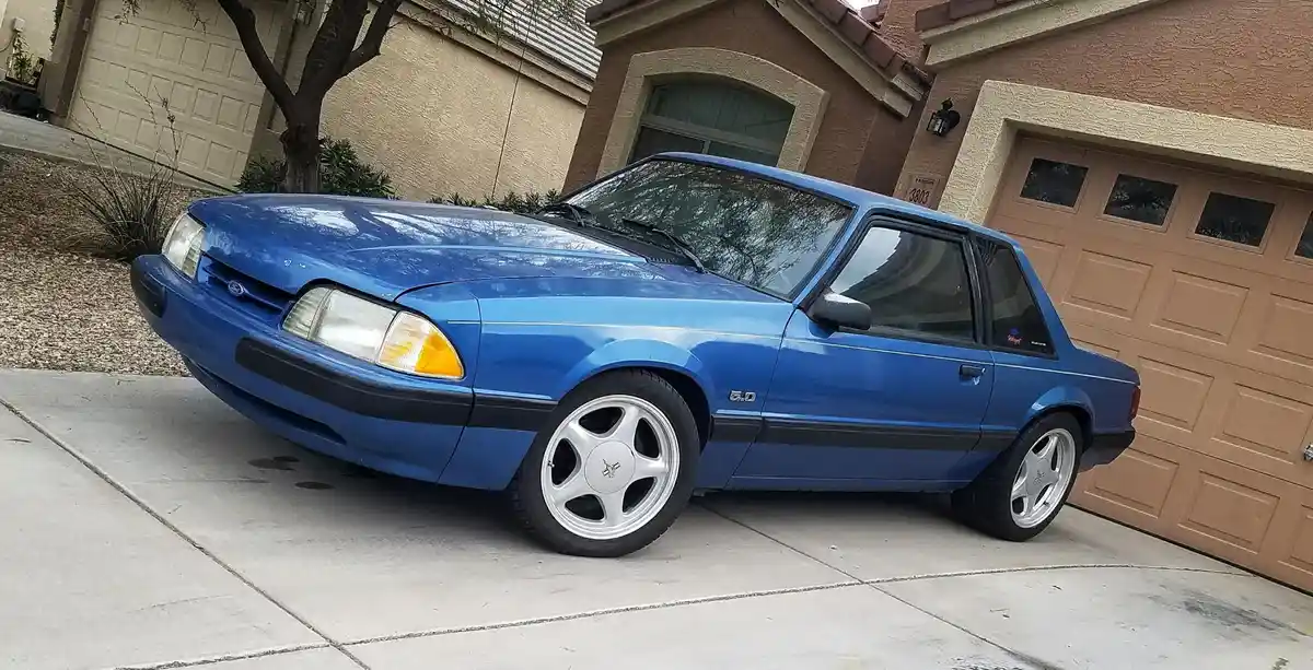 A blue 1993 Ford Mustang 5.0 is parked on a concrete driveway in front of a residential house with a tan garage door. The…