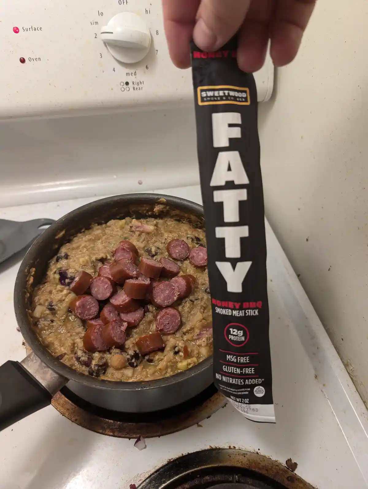 A hand holds a 'FATTY Honey BBQ Smoked Meat Stick' package over a pan of food on a stove. The pan contains a thick, chunky…
