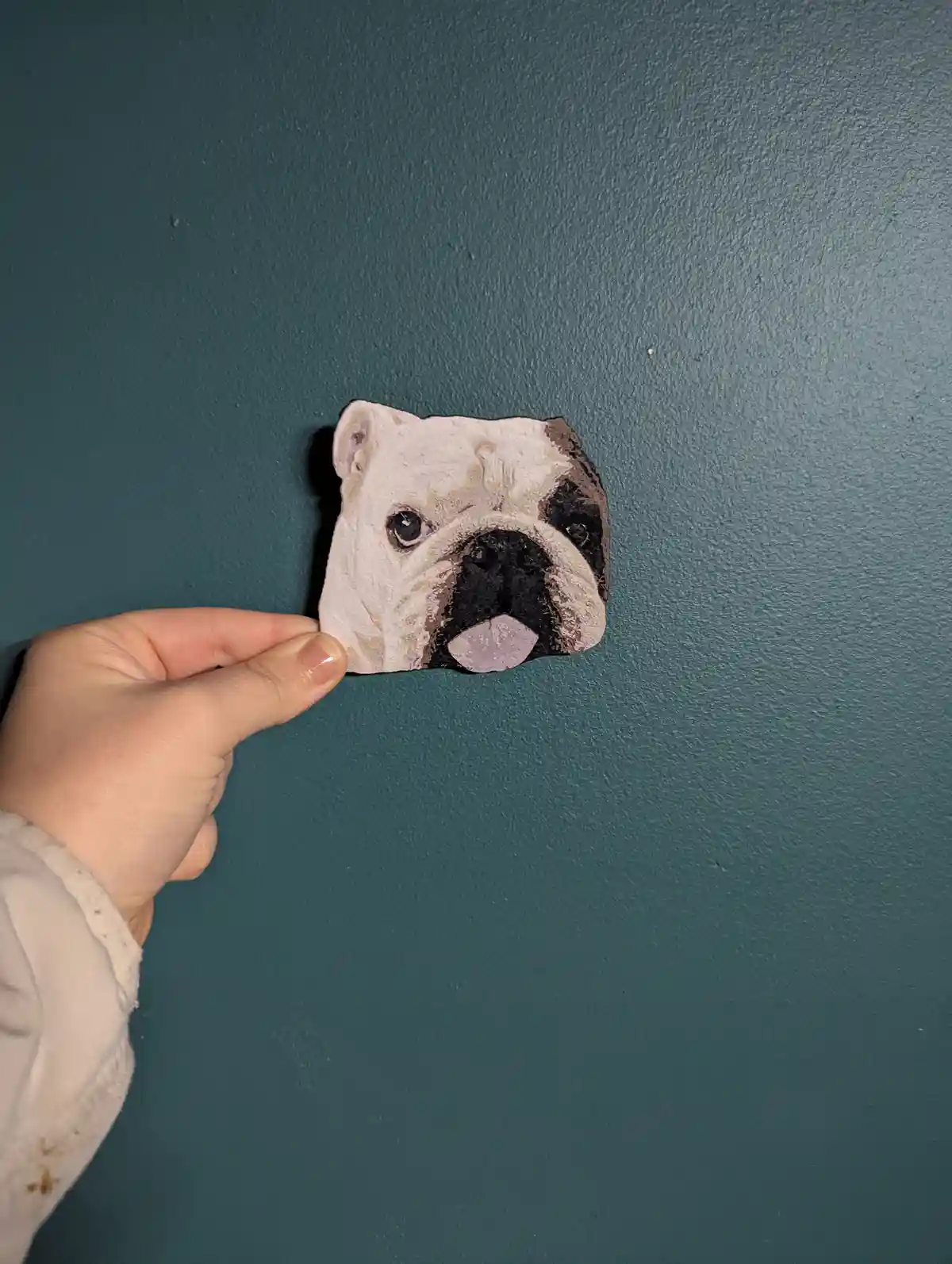 A hand holds up a small, roughly shaped clay sculpture of a bulldog's face against a teal wall. The sculpture has a…