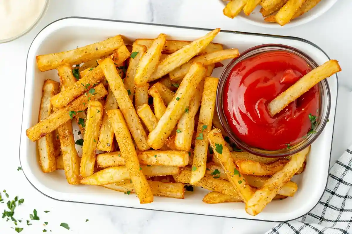 A white rectangular dish filled with golden-brown seasoned french fries, with a small glass bowl of red ketchup on the side…