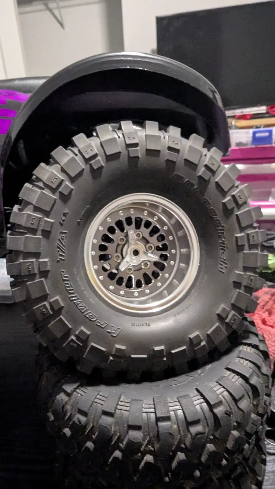 A close-up photo shows a large, rugged RC truck tire with a silver, multi-spoke wheel, stacked atop another tire. The tire…