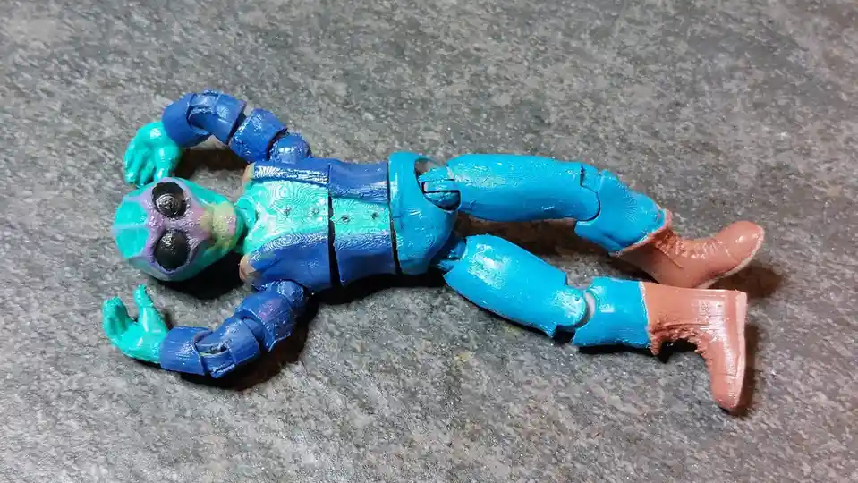 A 3D-printed action figure with a blue and teal body, purple and green face, and brown boots lies flat on a gray textured…