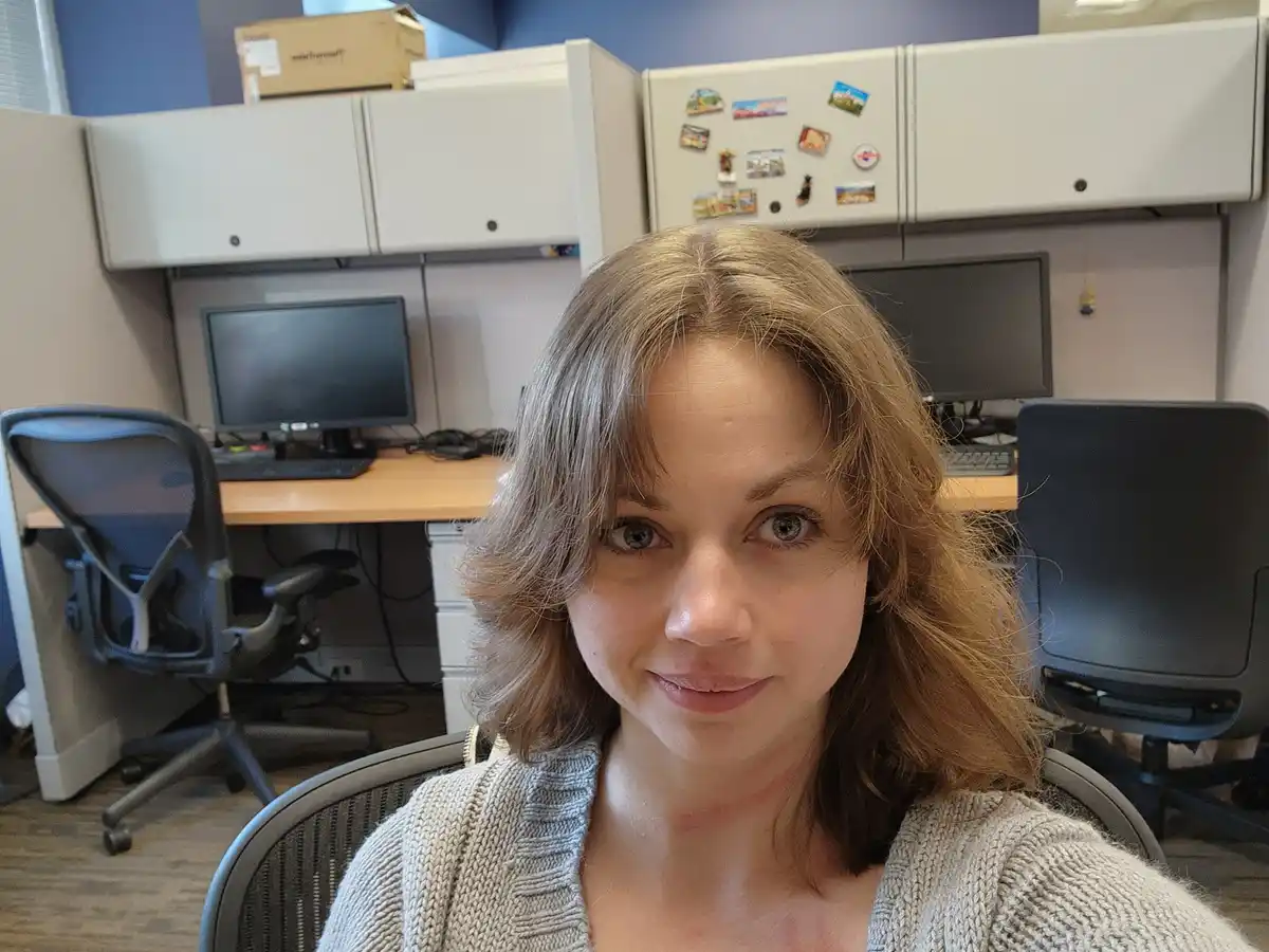 A woman with light brown hair and a neutral expression sits in an office cubicle, wearing a beige cardigan. Her neck and…