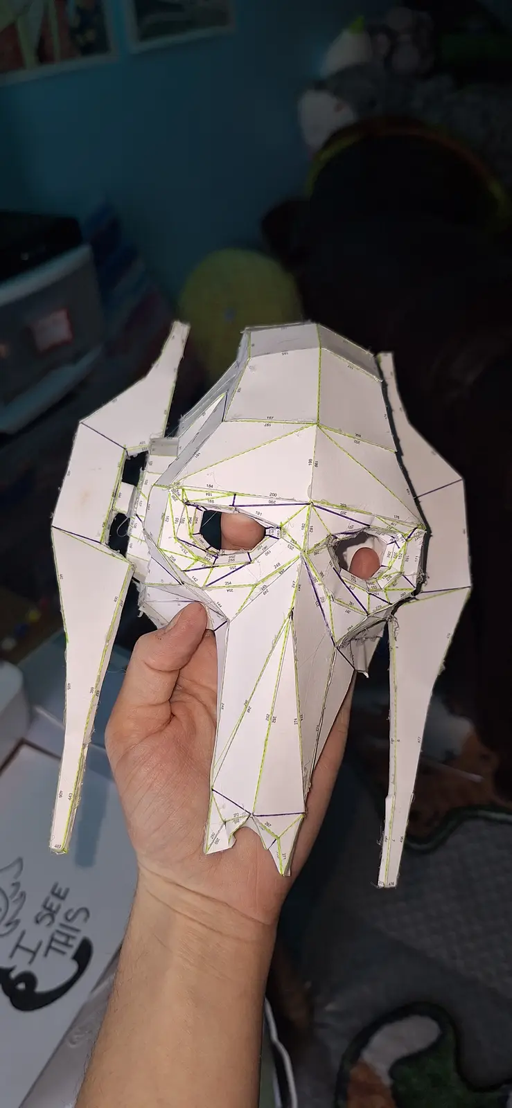 A hand holds a white paper mask with a geometric, angular design resembling a skull or alien head, featuring numerous…
