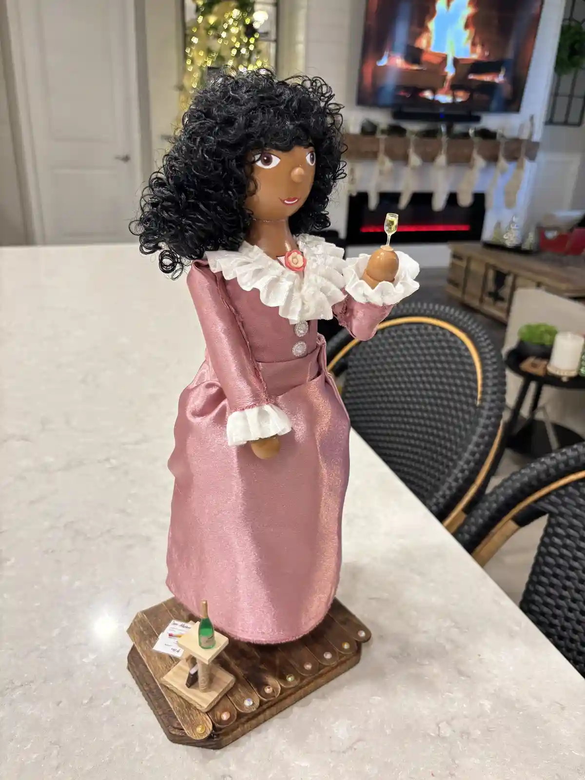 A decorative wooden doll with dark skin and curly black hair, wearing a pink satin dress with white ruffles, stands on a…