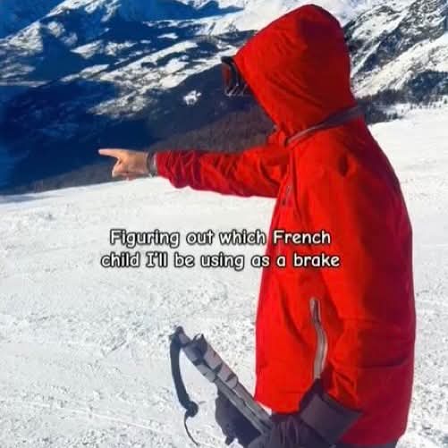 A person in a red hooded jacket stands on a snowy mountain slope, pointing toward the valley below while holding ski poles…