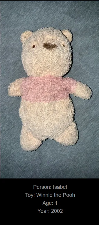A worn, light-colored plush Winnie the Pooh toy wearing a pink shirt lies on a gray fabric surface. Text overlaid at the…