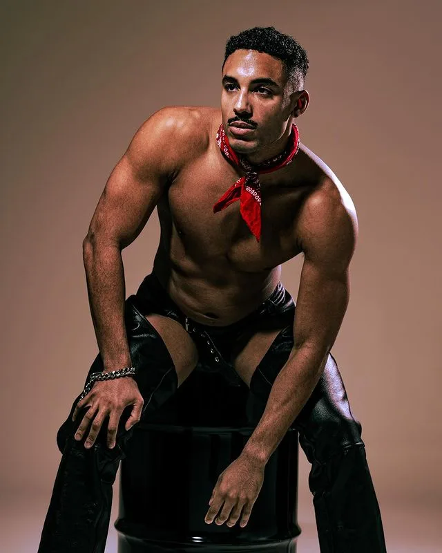 A shirtless, muscular man with a mustache and red bandana around his neck poses confidently while sitting on a black barrel…