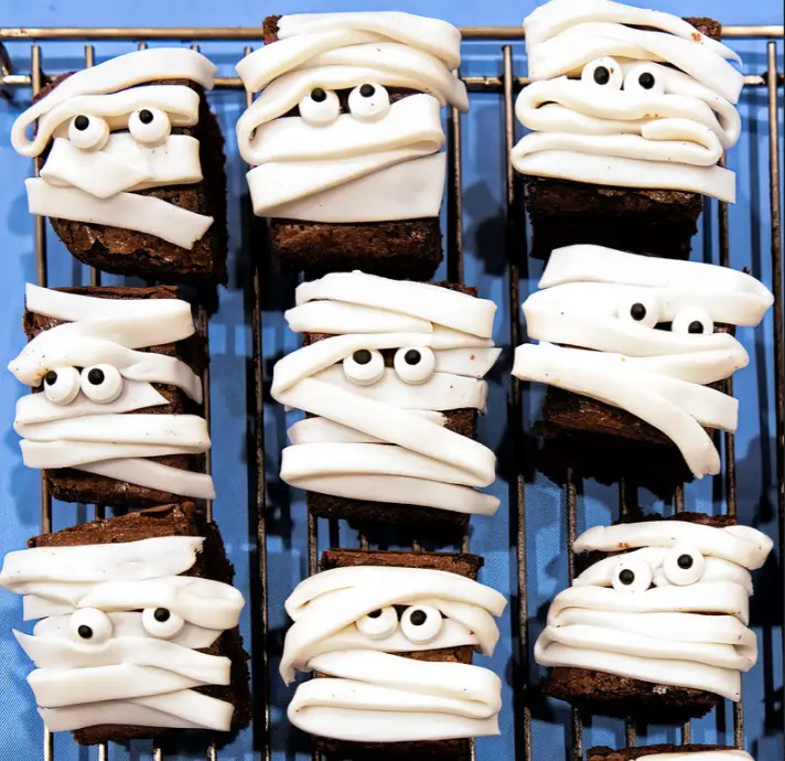 nine brownies decorated to look like mummies with white icing bandages and candy eyes, arranged on a wire cooling rack…