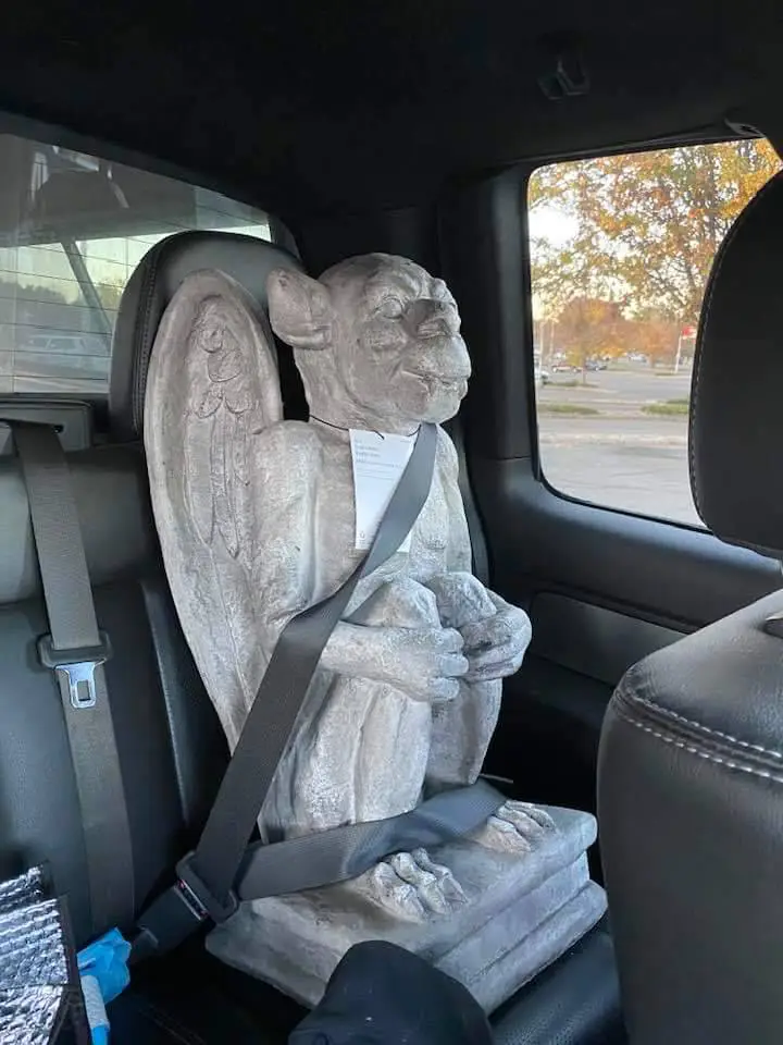 A stone gargoyle statue, with wings and a grimacing face, is strapped into a car seat with a seatbelt. The statue sits…
