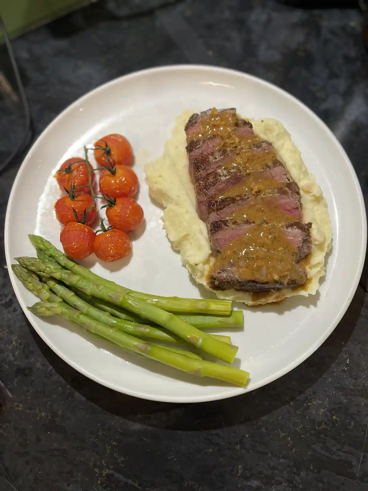 A white plate holds a gourmet-looking meal: sliced medium-rare steak with brown sauce over mashed potatoes, accompanied by…