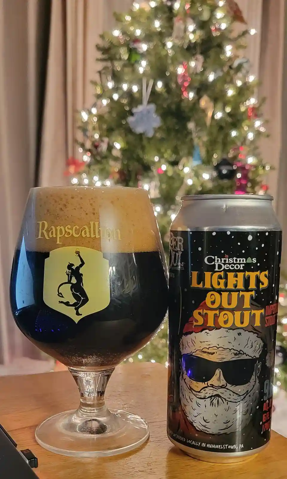 A dark beer in a 'Rapscaillon' branded glass sits next to a can labeled 'Christmas Decor Lights Out Stout' featuring a Santa…