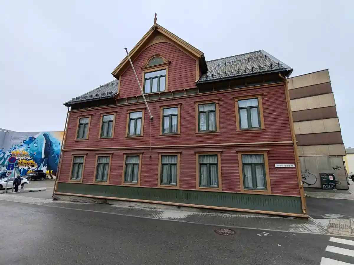 a red wooden building with green trim and multiple windows is visibly tilted at an angle, appearing to lean precariously…