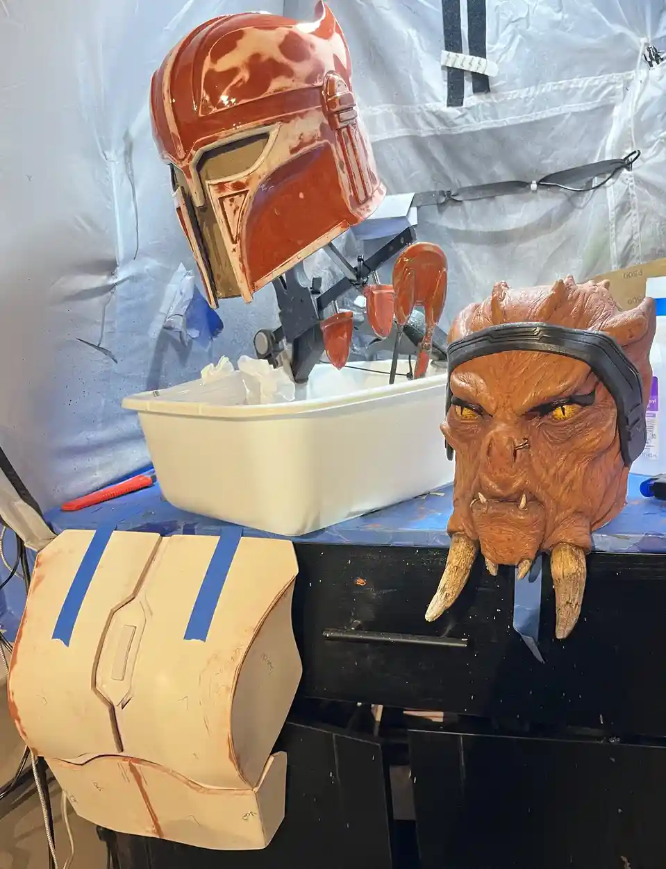A workshop scene shows three Star Wars-inspired costume pieces: a red Mandalorian helmet on a stand, a detailed orange alien…