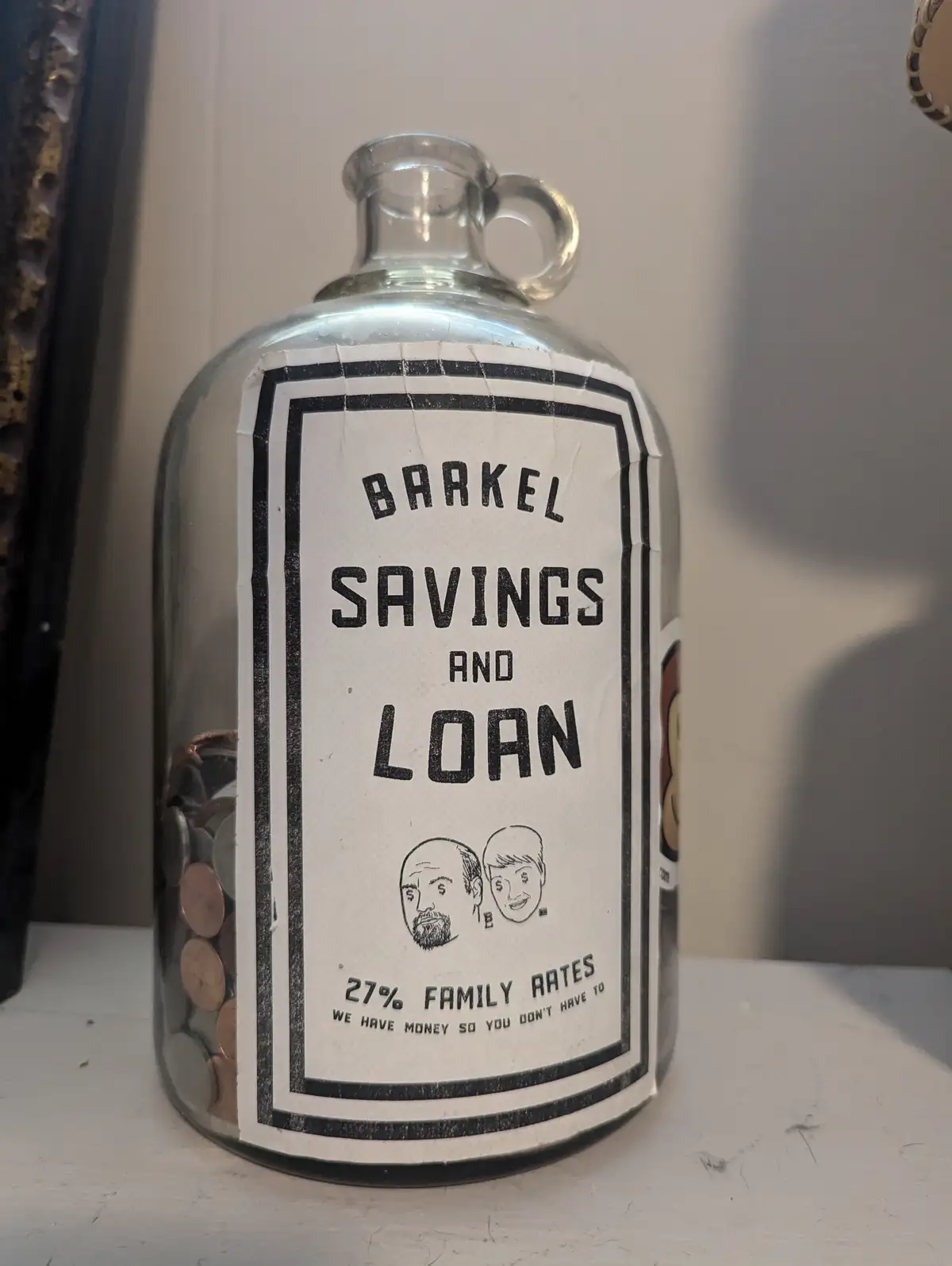 A clear glass bottle with a handle, filled with coins, has a handmade label that reads 'BARKEL SAVINGS AND LOAN', '27%…