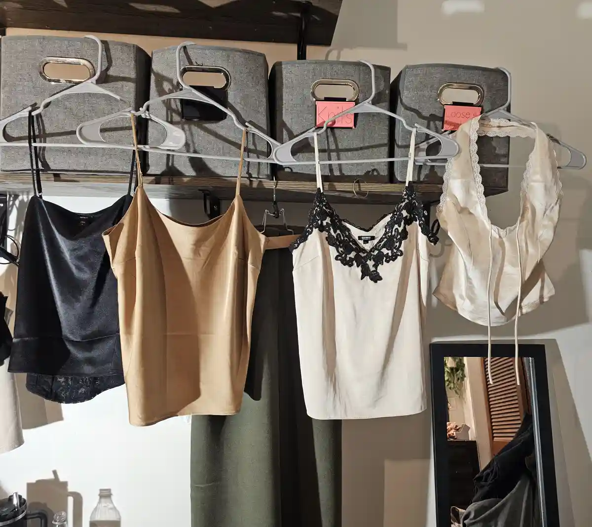 Four tops hang on white hangers from a wooden shelf, with gray fabric bins labeled 'Knits' and 'Loose Socks' above them. The…