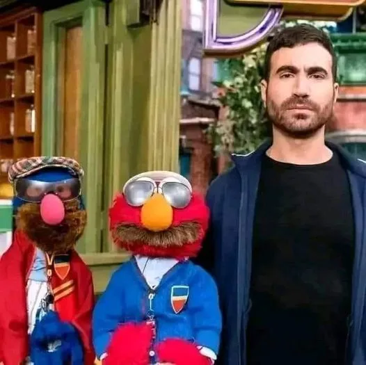 A man stands beside two puppets, Elmo and another Muppet, both wearing sunglasses and jackets with Italian flags. The man…