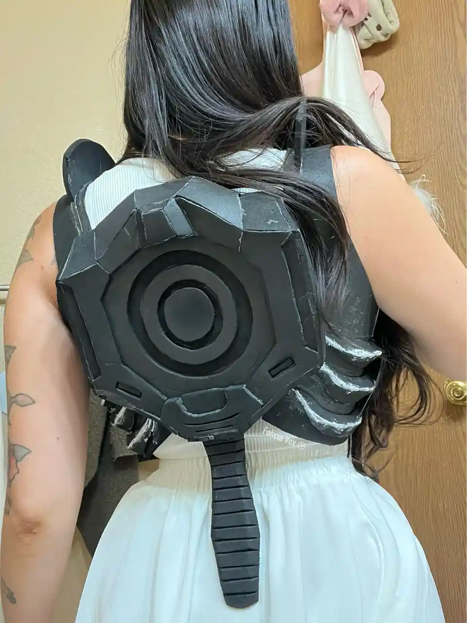 A person with long dark hair and a tattooed arm is seen from behind, wearing a large, black, angular, gear-shaped backpack…