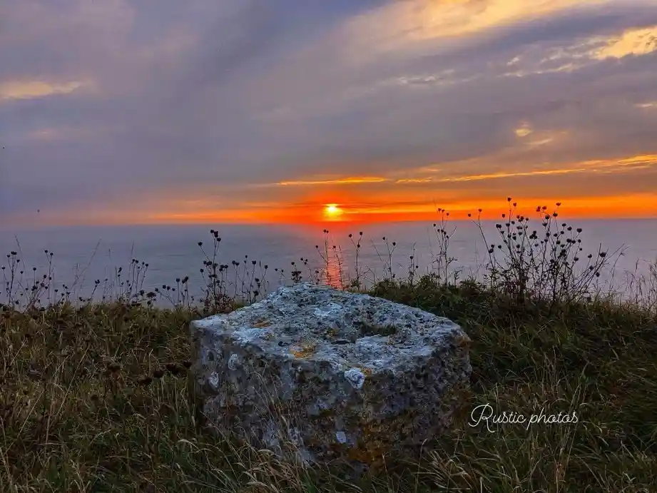 A scenic photograph of a sunset over the ocean, with a large, weathered stone in the foreground on a grassy cliff. The sky…