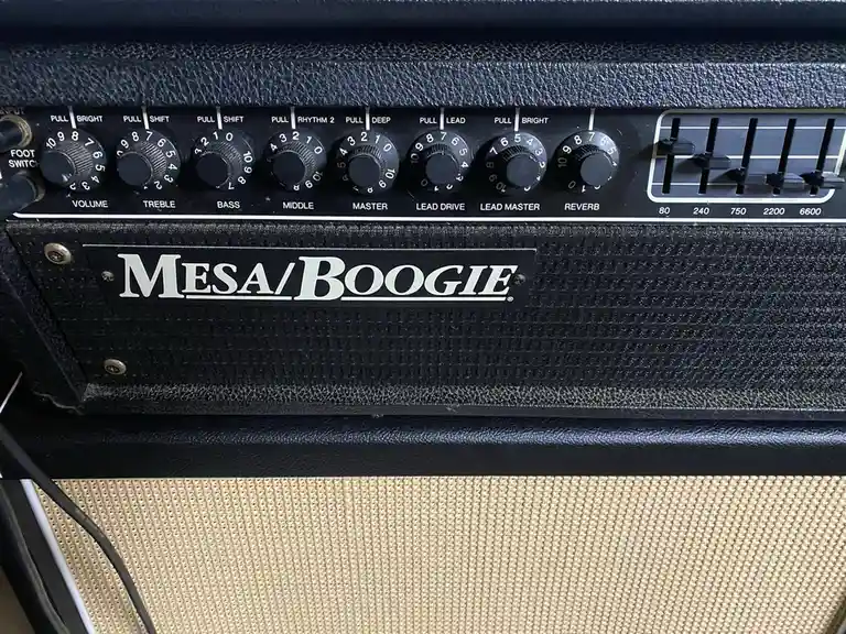 A close-up of a black Mesa/Boogie guitar amplifier head showing numerous knobs and switches labeled with functions like…