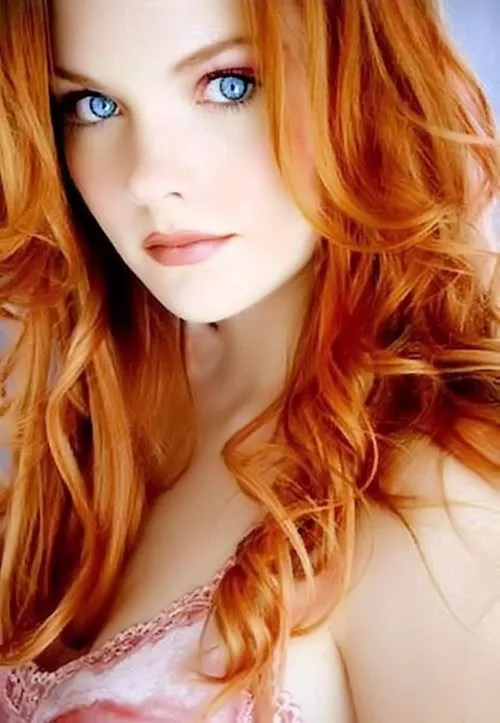 A close-up portrait of a woman with vibrant red, wavy hair and striking blue eyes, wearing a pink lace top. The image has…