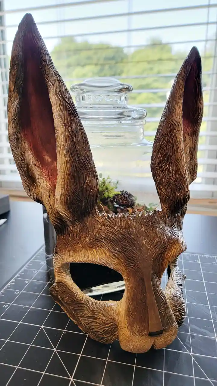 A wooden fox mask with large, detailed ears sits on a black gridded cutting mat, its eye sockets empty and the top opening…